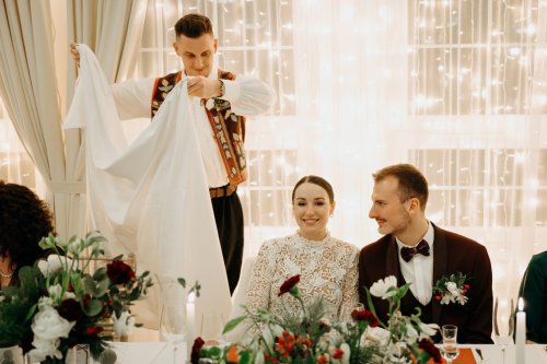 Bíž Starejší v ľudovom kroji počas svadobnej hostiny rozprestiera biely obrus pred usmiatym novomanželským párom.