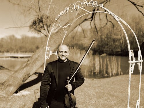 Elegantný huslista z projektu Bridgerton Violin Experience pózuje s husľami pod bielou ozdobnou bránou v prírodnom prostredí pri jazere.