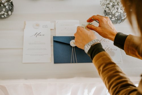 Detailný záber na elegantné svadobné oznámenia, menu a modrú obálku s pečaťou, ktoré aranžujú ruky fotografky.