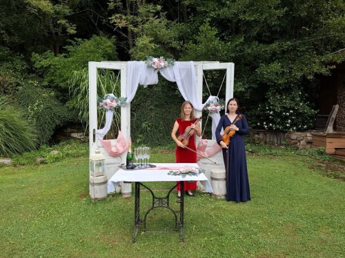 Dve huslistky v elegantných šatách pózujú pri zdobenej svadobnej bráne v záhrade, zachytené v rámci série Naše foto zo svateb.