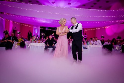 Mladomanželia tancujú svoj prvý svadobný tanec v oblakoch nízkej hmly v sále s fialovým nasvietením.