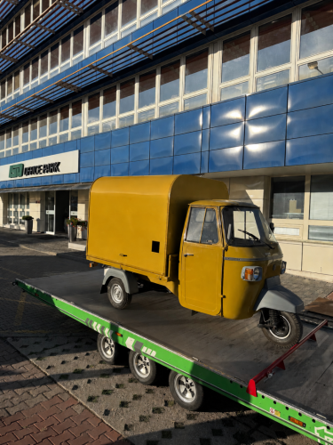 Žltá trojkolka Piaggio Ape, základ pre mobilný svadobný bar Prosecco Piškot, naložená na prepravnom prívese pred budovou.