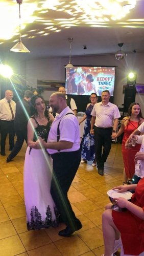 Svadobčania tancujúci redový tanec pod vedením DJa a moderátora v osvetlenej sále.