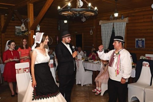 Starejší s gitarou v slovenskom ľudovom kroji počas svadobnej zábavy s nevestou a ženíchom v kolibe.