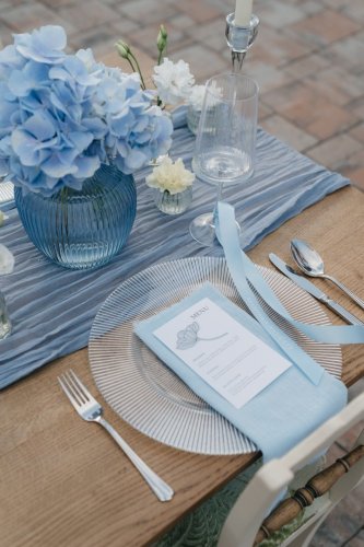 Elegantné svadobné menu na mieru od LU.C design položené na svetlomodrom obrúsku a sklenenom tanieri s modrou kvetinovou výzdobou.