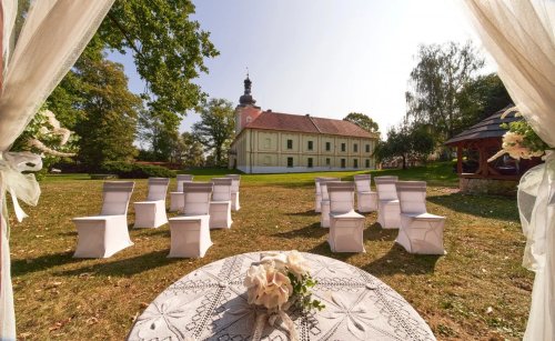 Svadobný obrad v Lešany Garden s bielymi stoličkami na trávniku pred historickým kaštieľom za slnečného dňa.