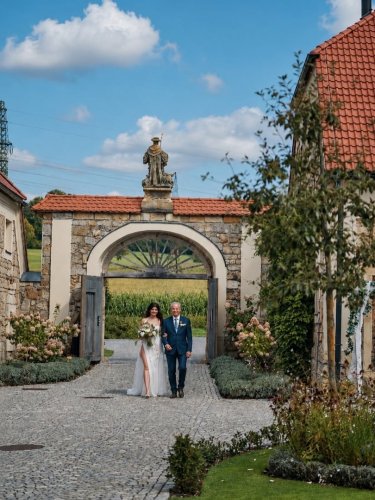 Nevesta v bielych svadobných šatách kráča pod ruku s otcom cez historickú kamennú bránu v areáli Tvrz KRASSA.